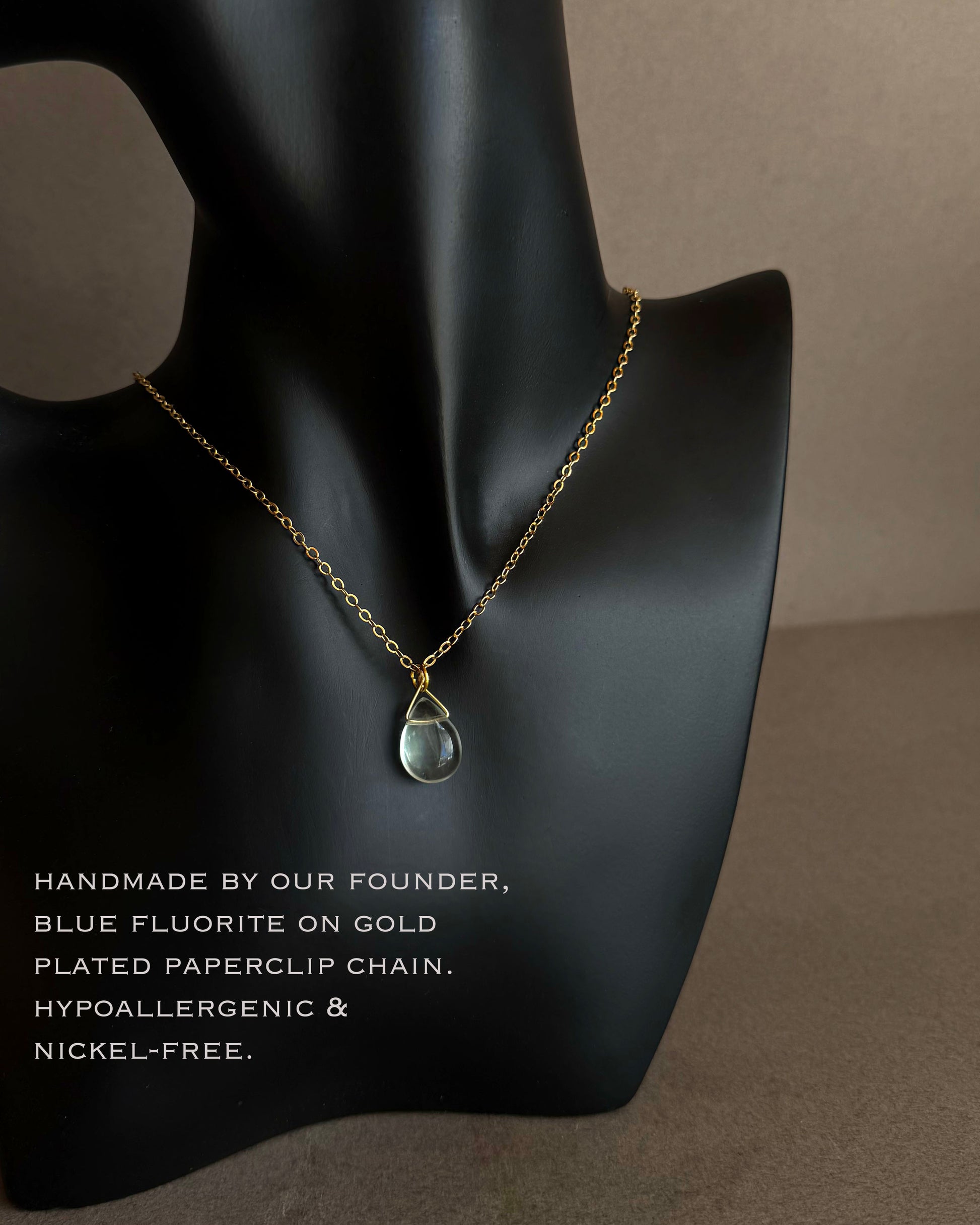 Necklace with a teardrop pendant on a black mannequin bust with product details. Gold paperclip chain necklace with blue fluorite teardrop pendant detail
