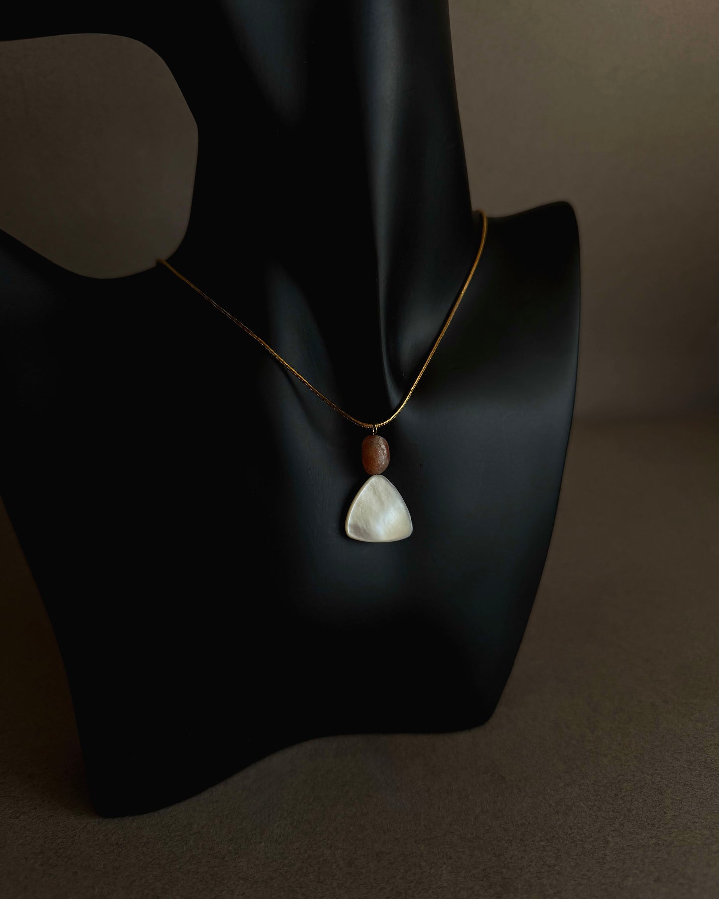 Necklace with a pendant on a black mannequin bust against a dark background Gold waterproof snake chain necklace with mother of pearl and sunstone pendant on textured background
