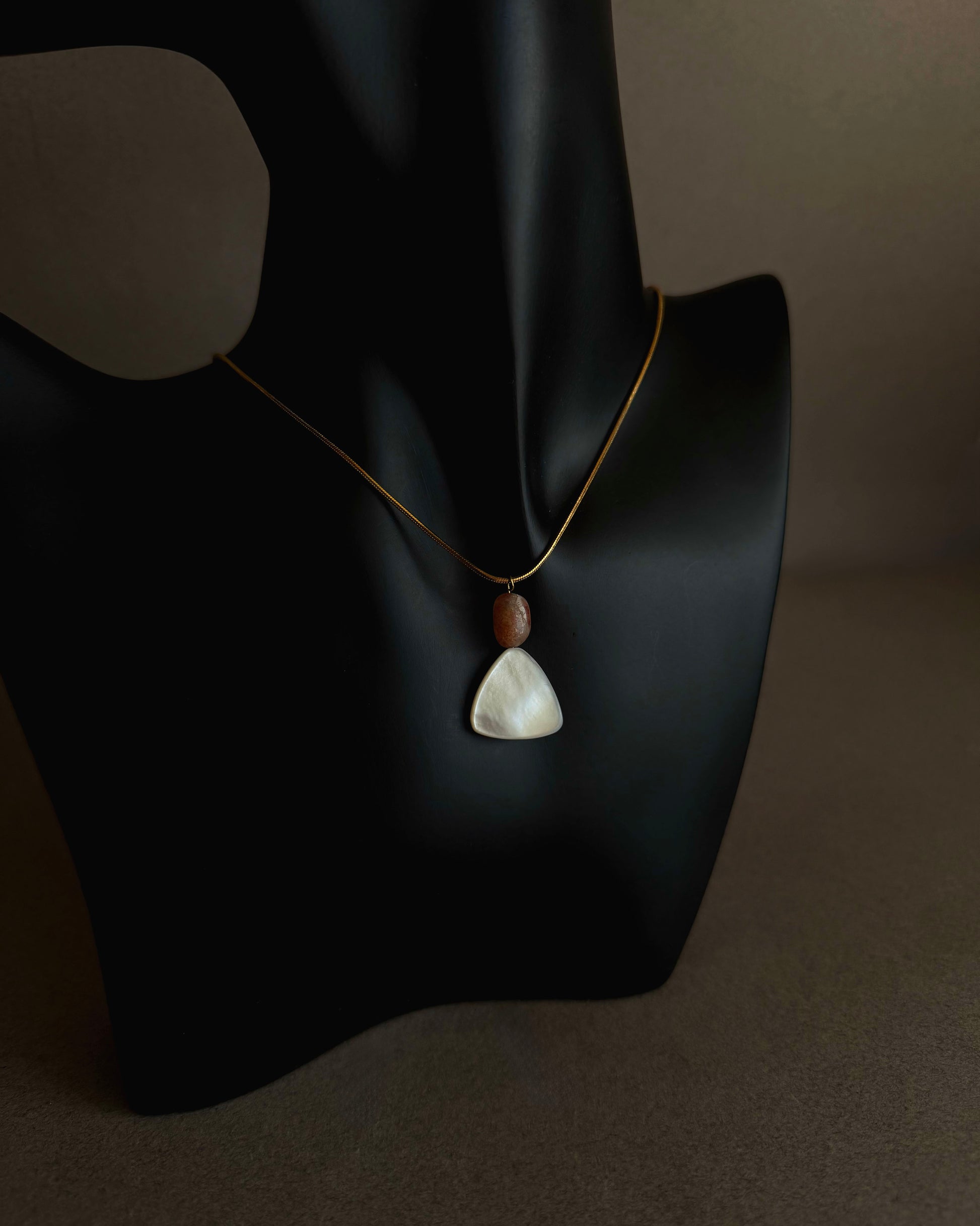 Necklace with a pendant on a black mannequin bust against a dark background Gold waterproof snake chain necklace with mother of pearl and sunstone pendant on textured background

