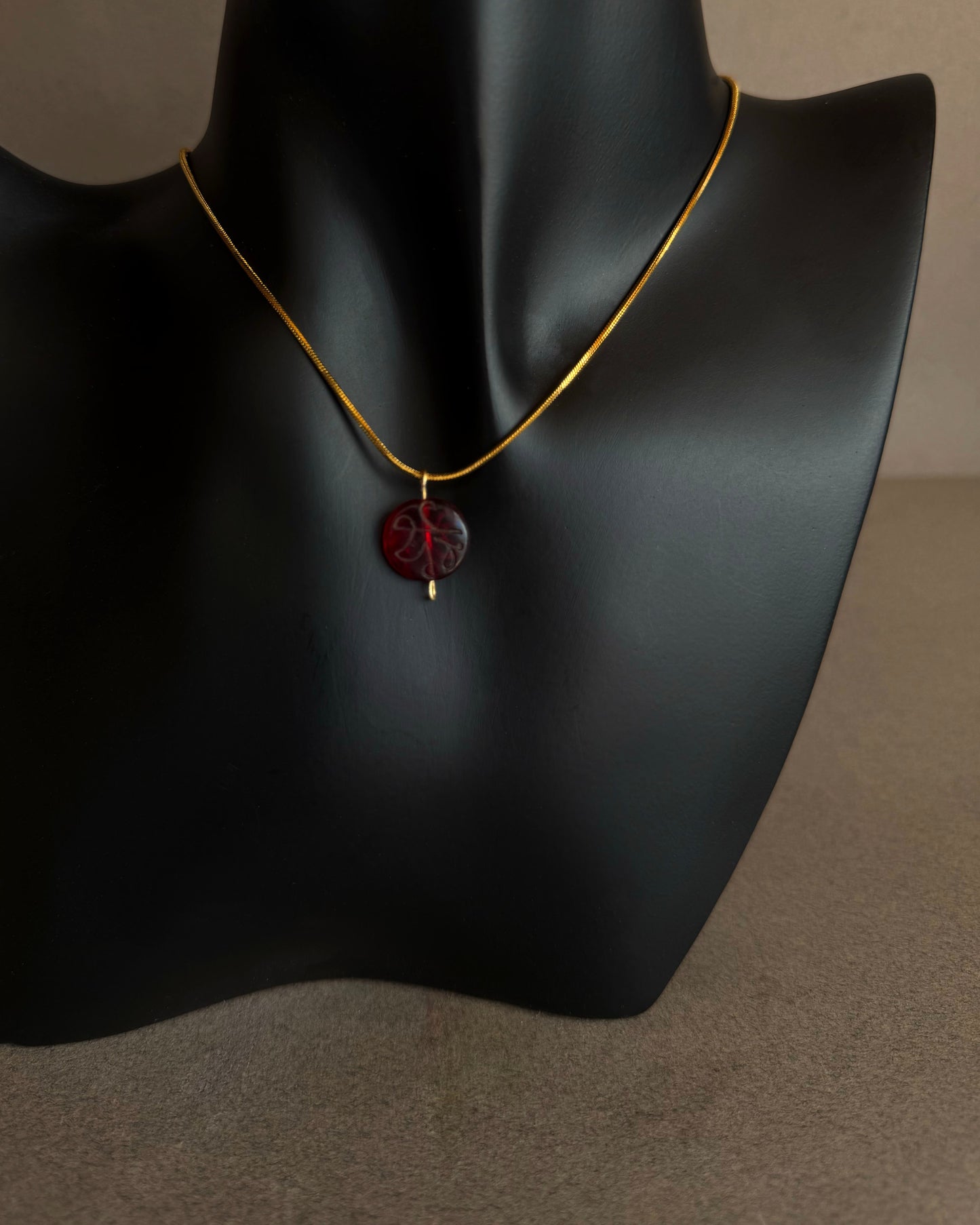 Necklace with a red pendant on a black mannequin bust. Gold waterproof snake chain necklace with round red garnet pendant on textured background
