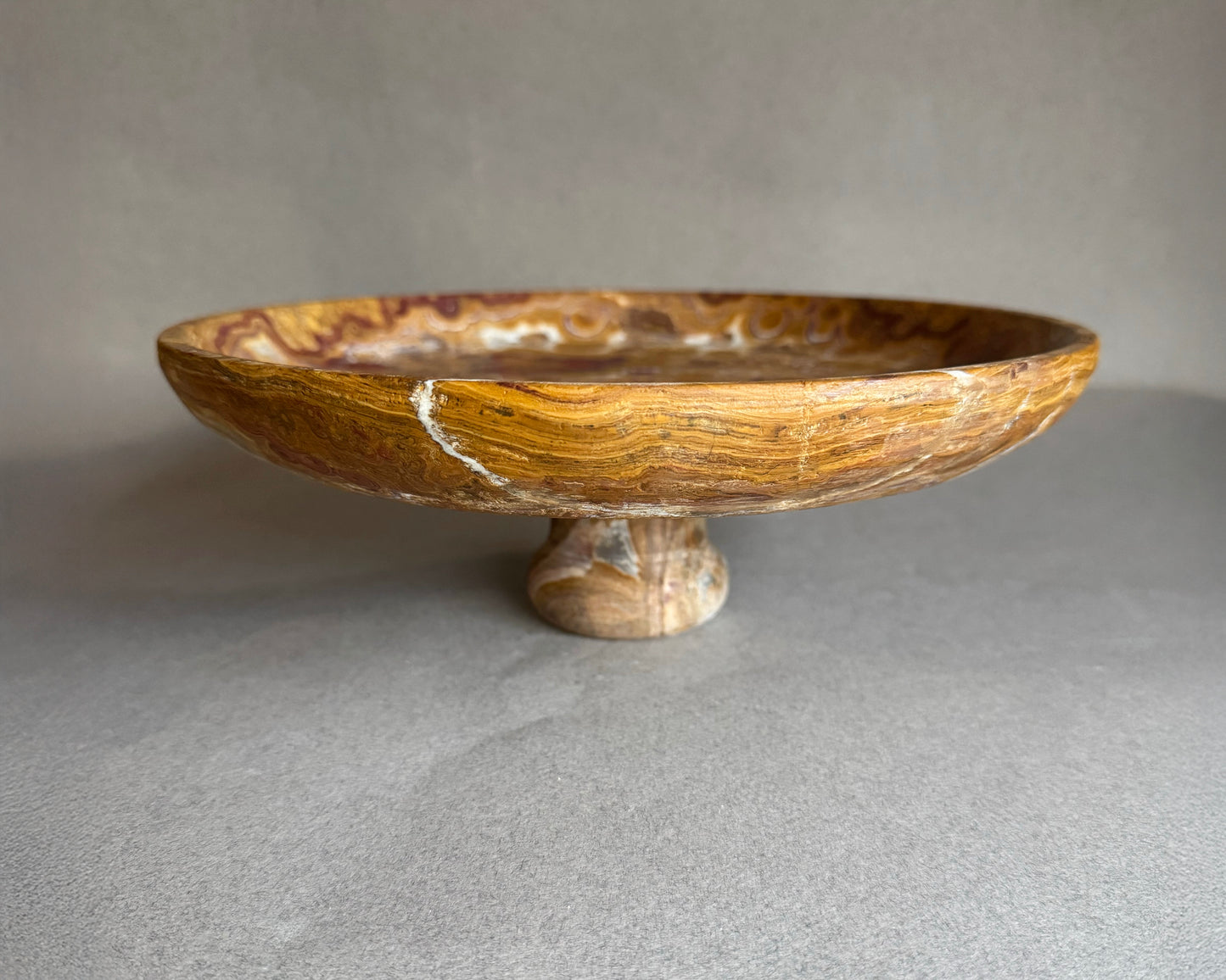 Hand-carved brown onyx pedestal bowl with swirling amber and rust veining, styled as a natural stone fruit bowl centerpiece.