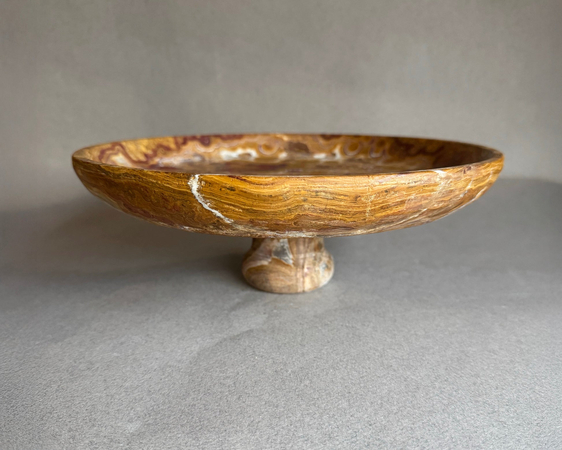 Hand-carved brown onyx pedestal bowl with swirling amber and rust veining, styled as a natural stone fruit bowl centerpiece.