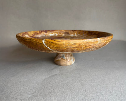 Hand-carved brown onyx pedestal bowl with swirling amber and rust veining, styled as a natural stone fruit bowl centerpiece.