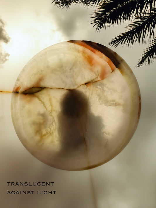 Translucent sphere with a dark spot against a light background, palm leaves visible. Top view of natural onyx pedestal bowl featuring creamy ivory stone with soft amber, green, and blush mineral veining
