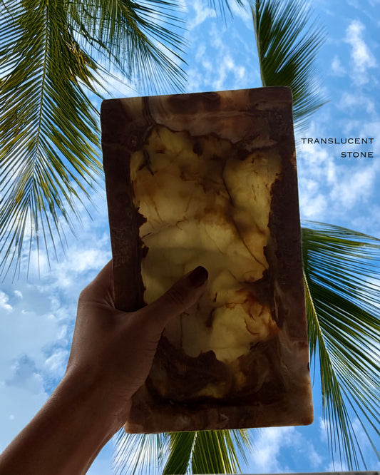 Hand holding a translucent stone with palm trees and blue sky in the background Rectangular natural onyx catchall tray with layered amber, ivory, and caramel veining photographed at an angle
