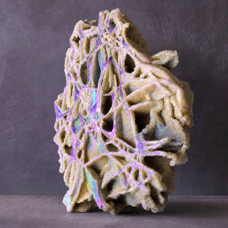 An organic-shaped decorative object sphalerite slab sculpture with a textured, coral-like surface in warm beige and iridescent lavender-purple tones, standing upright against a dark gray studio background.