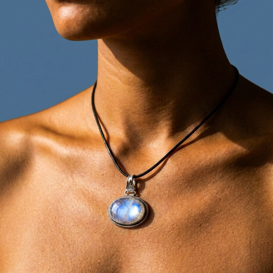 A close-up lifestyle shot of a model wearing a black cord necklace with a rainbow moonstone pendant silver bezel containing a glowing, light-blue moonstone pendant.