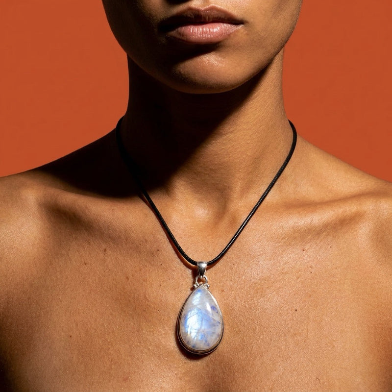 A close-up of a model's neck and chest wearing a black cord necklace with a teardrop-shaped iridescent RAINBOW MOONSTONE pendant on a warm orange background.