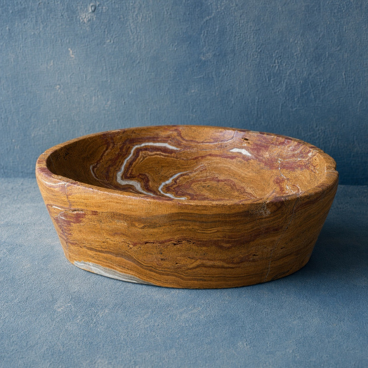 A round onyx stone bowl with a rich golden-brown and reddish-brown wood grain pattern, set against a textured blue-gray background.