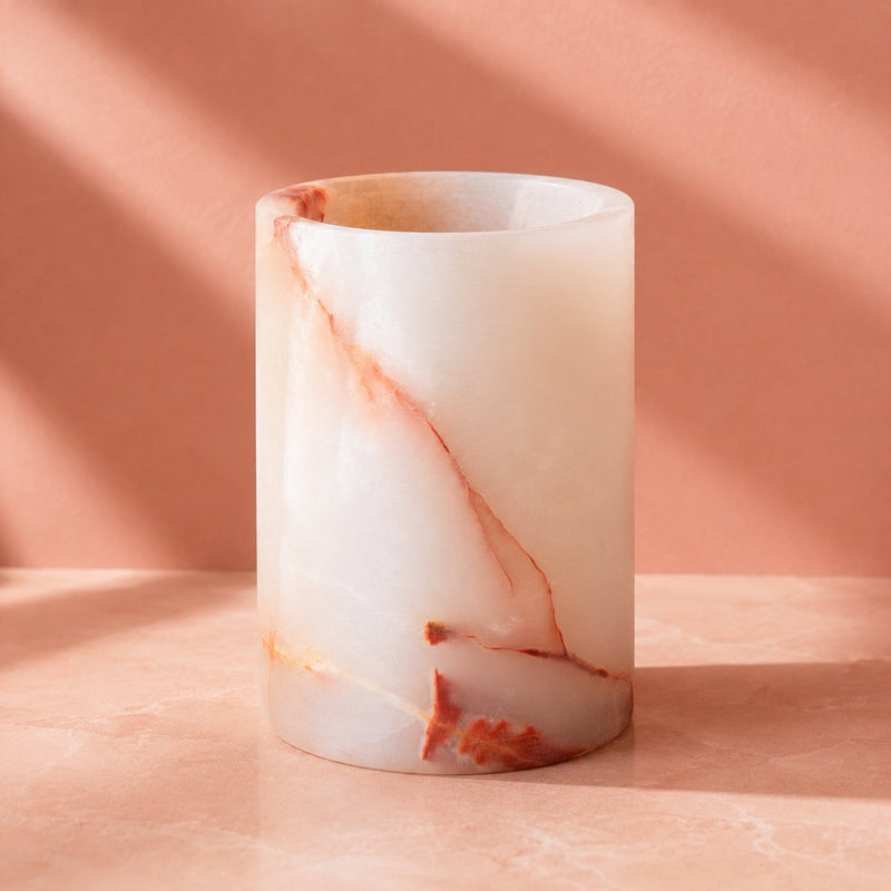 A cylindrical onyx stone vessel made of milky white onyx with elegant terracotta and orange veining, set on a matching peach-colored surface with soft diagonal shadows.