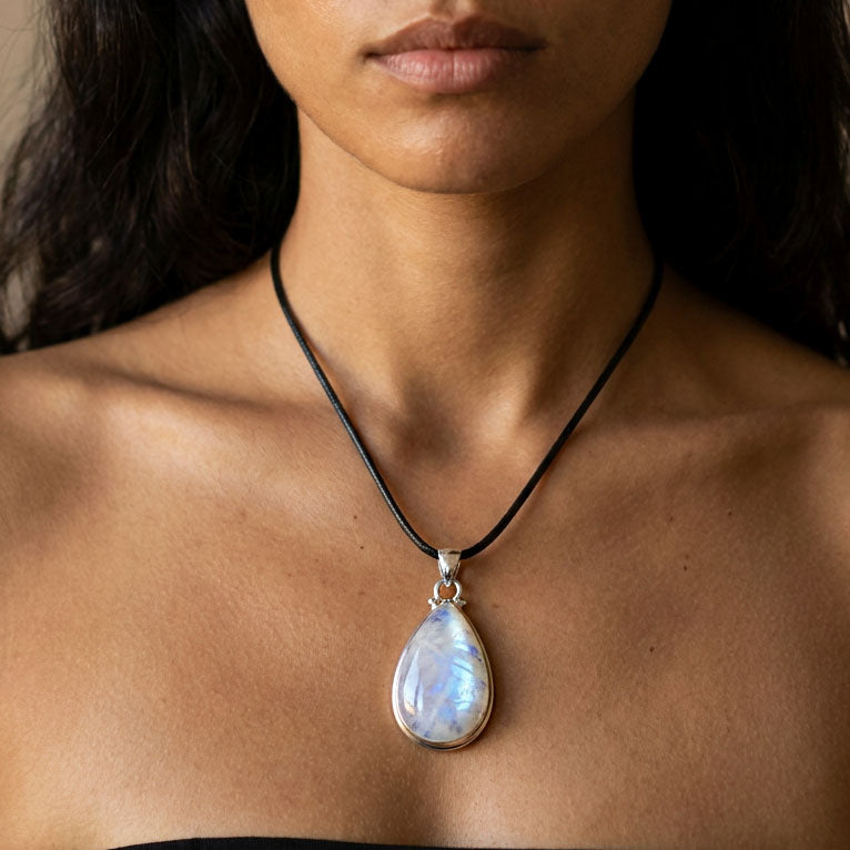 A close-up of a woman wearing a teardrop-shaped iridescent moonstone pendant necklace on a black cord with a silver bezel.