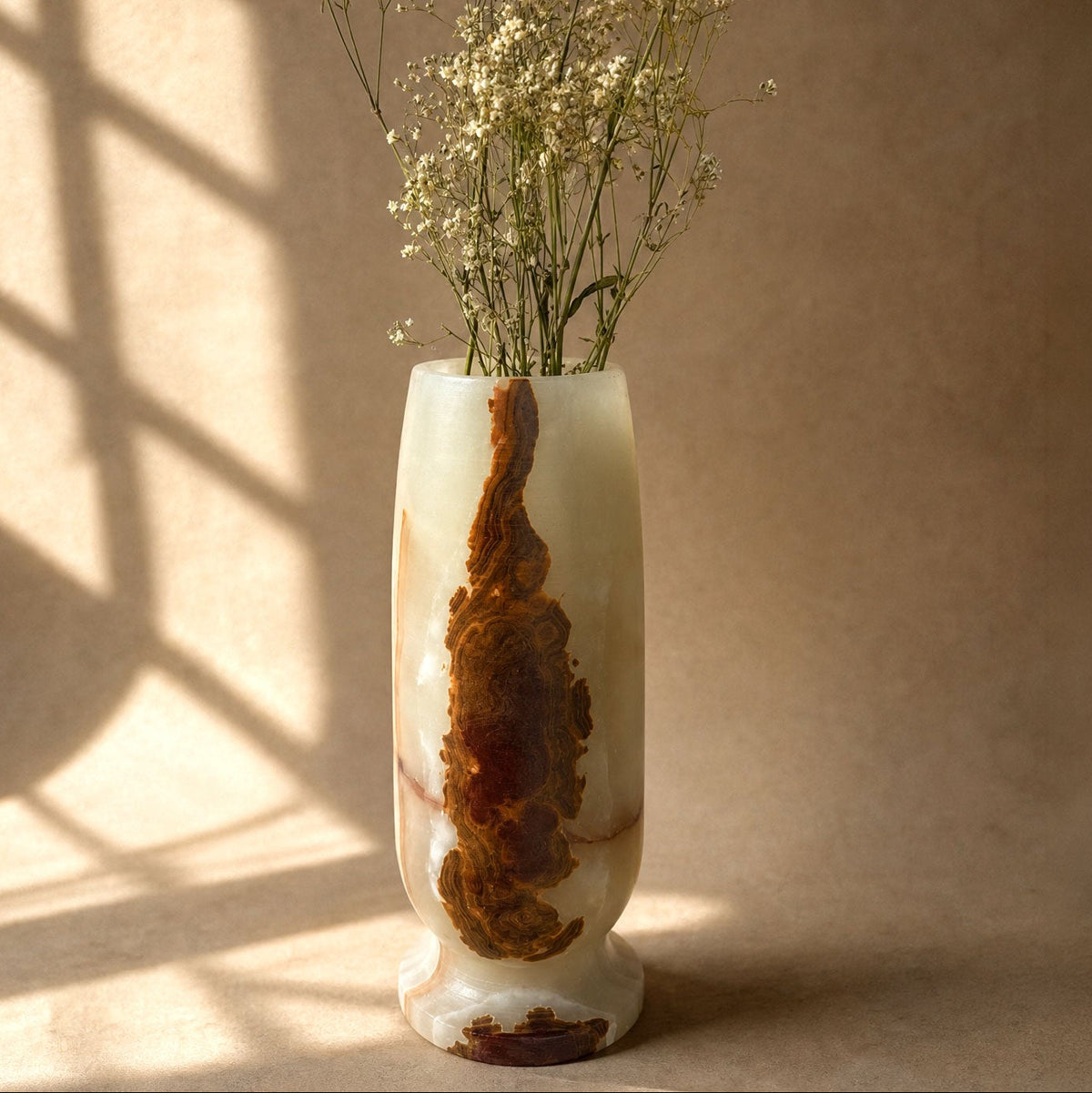 A tall, cylindrical carved stone vase with a natural brown and cream marbled pattern, filled with dried white baby's breath flowers, set against a warm beige background with soft window shadow patterns.