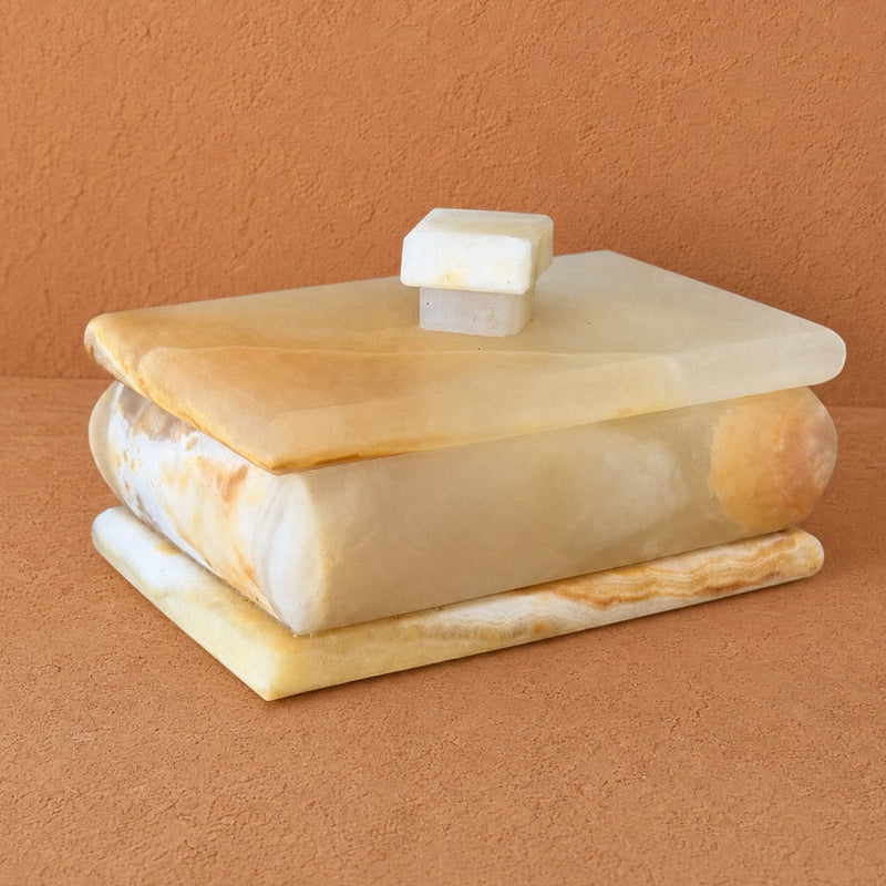 A rectangular peach yellow onyx stone box with a matching lid and small top knob, photographed against a warm terracotta textured background. The stone has creamy off-white and soft tan marbling.