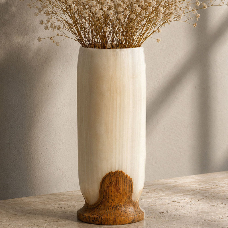A tall cylindrical ceramic vase with a cream-colored body and a natural brown wood-grain base, filled with dried baby's breath flowers, set on a stone surface against a textured wall with soft window shadows.
