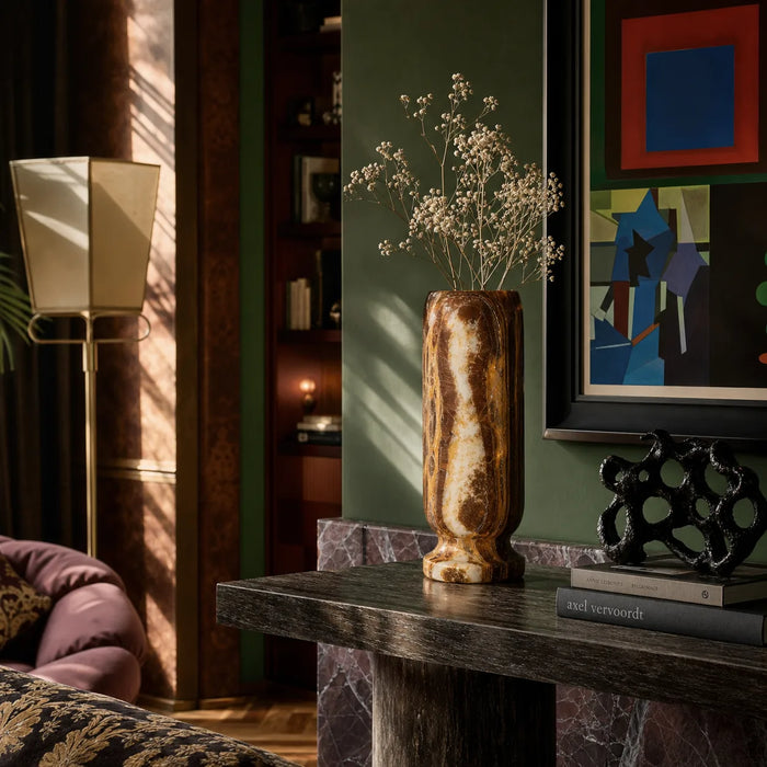 A tall, textured brown and gold marble-look vase filled with delicate white dried flowers, displayed on a dark marble-topped console table in a styled living room setting.