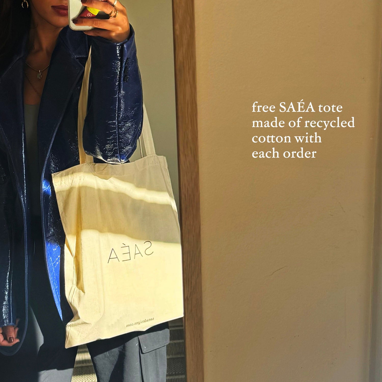 Person holding a beige tote bag with 'SAÉA' branding, standing in front of a neutral wall.