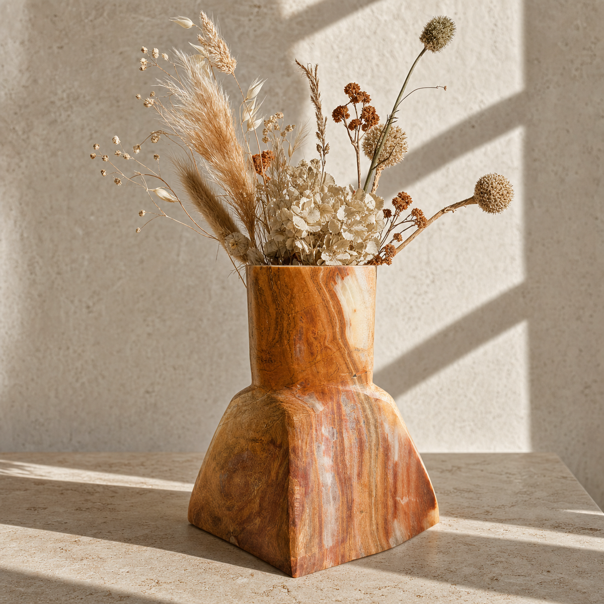 A sculptural terracotta onyx stone clay vase with a flared base, filled with dried botanicals, set on a stone surface against a textured wall with soft window shadows.