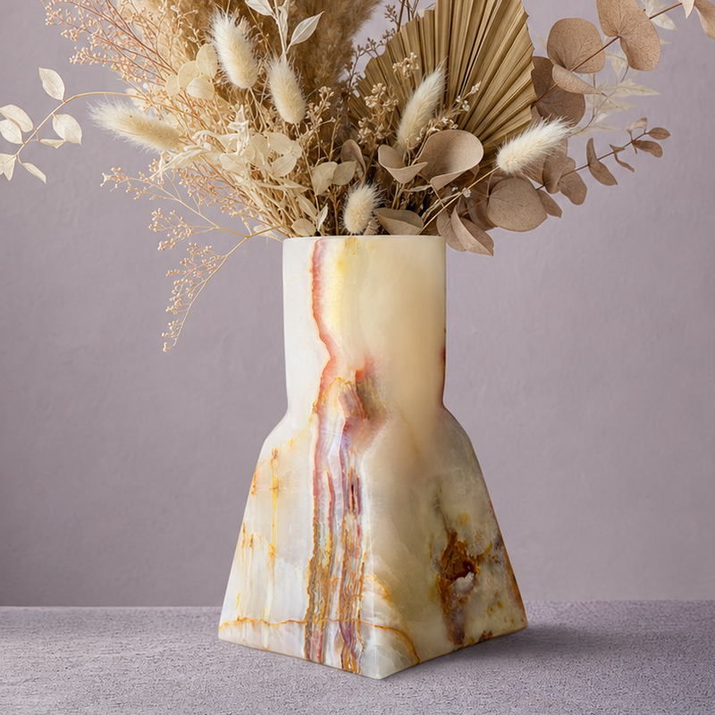 A sculptural onyx-style vase with cream, beige, and brown marbling, filled with a dried floral arrangement of pampas grass, eucalyptus, and palm leaves, set against a muted mauve-gray background.