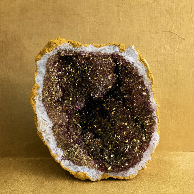 A natural geode specimen with a dark purple interior filled with gold glitter crystals, surrounded by a white crystalline layer and a golden-yellow outer border, set against a textured gold background.