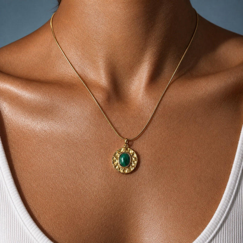 A gold chain necklace with a circular pendant featuring a textured gold border and a central green cabochon stone, shown worn on a model's neck.