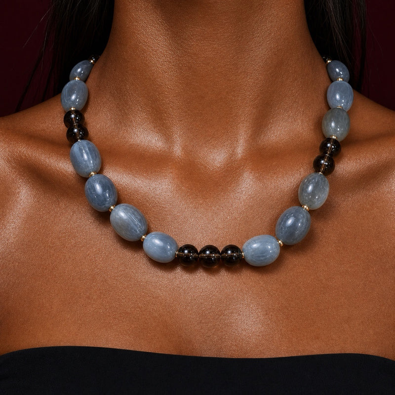 A close-up of a beaded necklace worn on a model's neck. The necklace features large, polished light blue oval beads separated by small gold spacers, with a section of smaller dark brown and black beads.