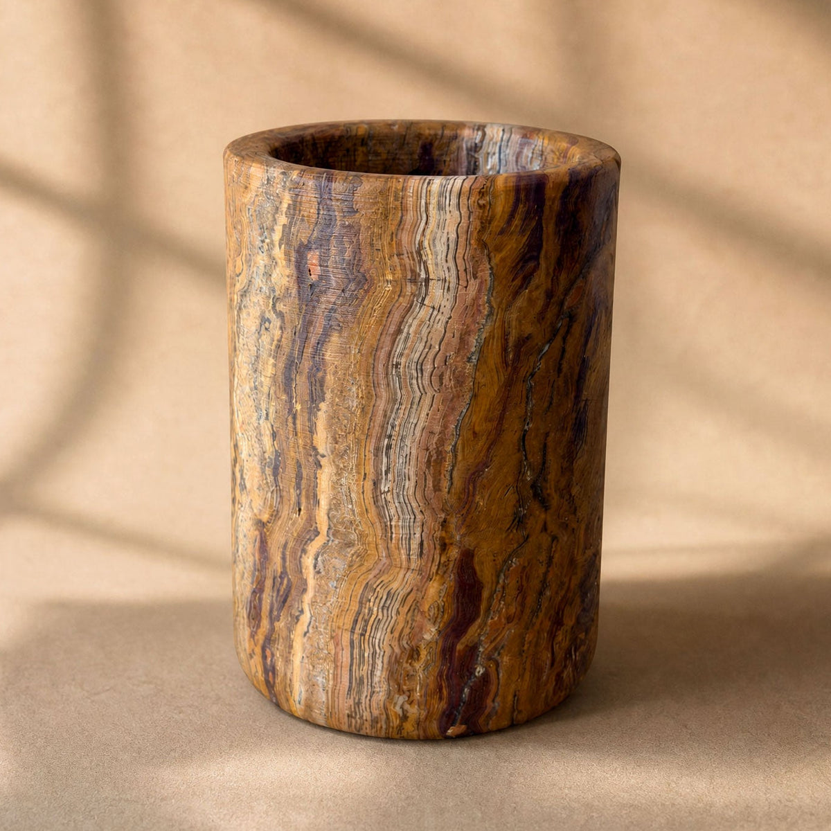 A cylindrical onyx cup or vase made from polished stone with rich brown, gold, and black wavy grain patterns, set against a warm beige background with soft window shadow overlays.