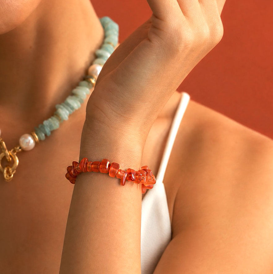 A close-up of a person wearing a chunky orange-red beaded bracelet on their wrist, with a light blue and white beaded necklace also visible against a warm terracotta background.