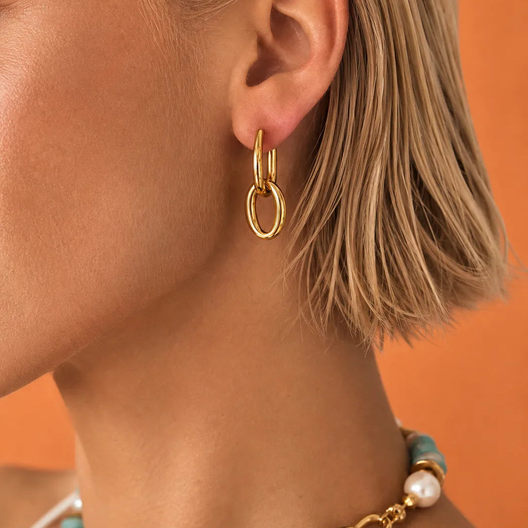 A close-up of a model's ear wearing a pair of gold hoop earrings, with a matching gold chain necklace partially visible. The model has short blonde hair and is set against a solid orange background.