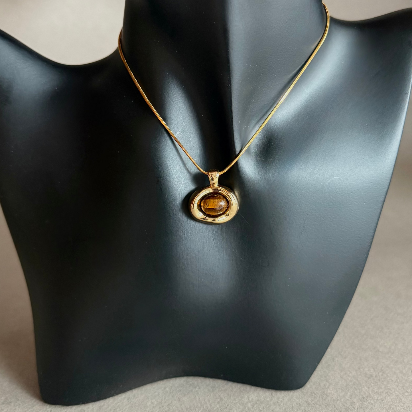Gold snake chain necklace with a amber tiger's eye pendant on a black mannequin bust.
