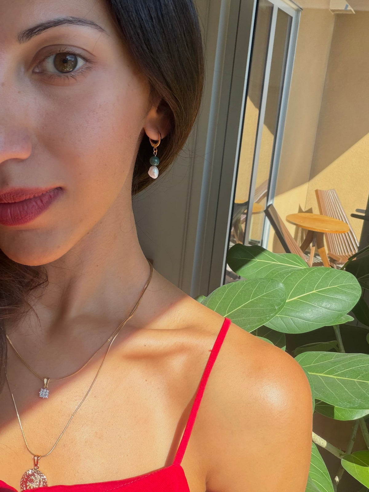 Woman wearing a red top with a blurred indoor background with gold filled huggie earrings with freshwater pearl and jade