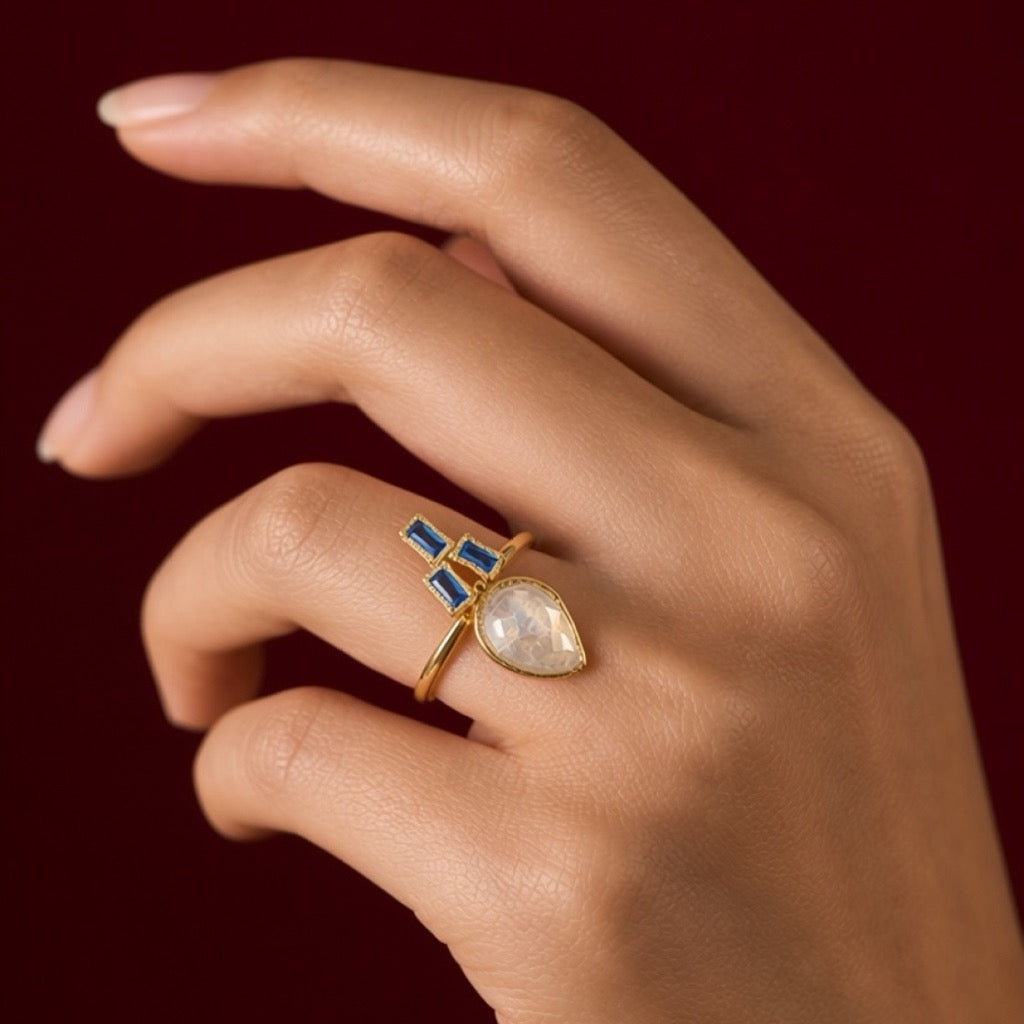 A gold ring with a pear-cut milky white stone and small blue rectangular stones, worn on a hand against a dark red background.