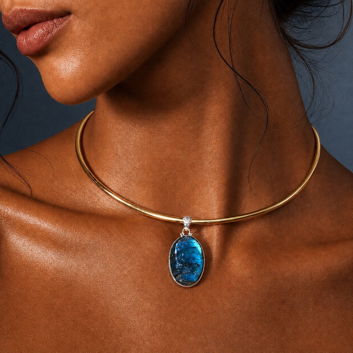 A close-up of a model wearing a gold collar necklace with a blue labradorite oval pendant in sterling silver and a small clear accent stone.