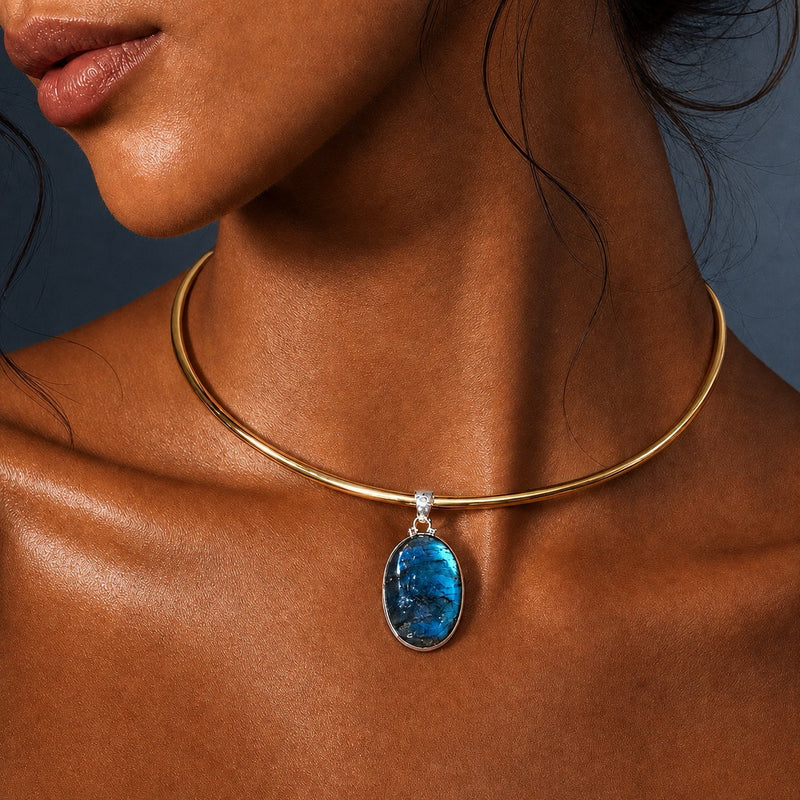 A close-up of a model wearing a gold collar necklace with a blue labradorite oval pendant in sterling silver and a small clear accent stone.