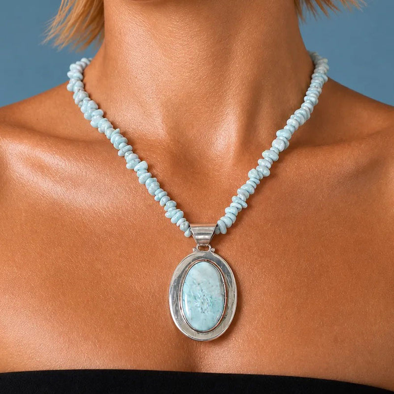 Oval larimar cabochon banded pale blue and white in a heavy sterling bezel, hung on a larimar strand and worn at mid-chest against a blue backdrop.