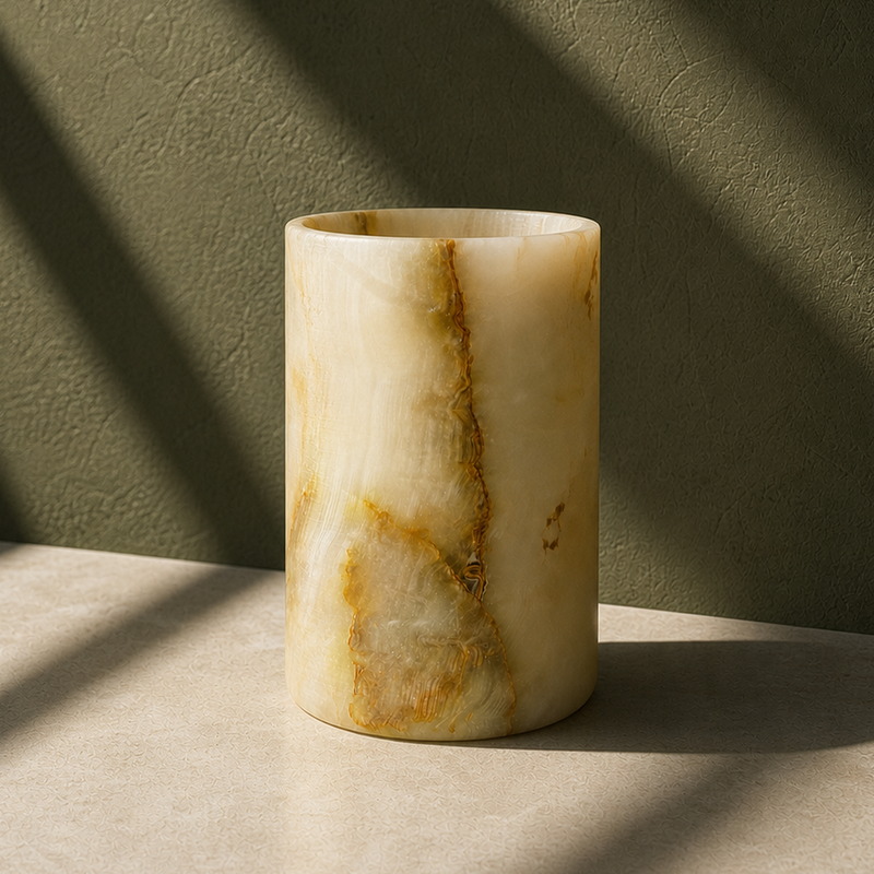 A cylindrical stone vase made of cream and tan marbled onyx or alabaster, standing on a light stone surface with dramatic diagonal shadows cast across a textured olive-green wall.