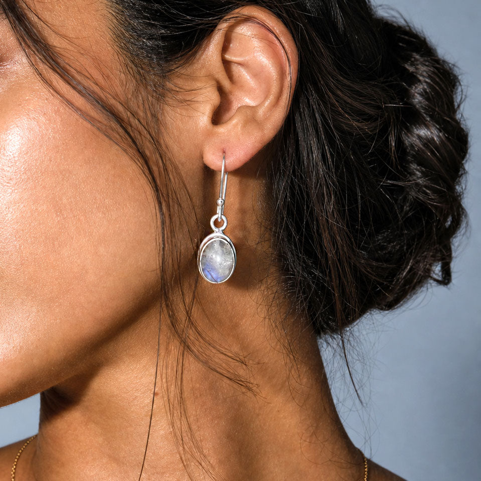 A close-up of a woman's ear wearing a silver dangle earring with a teardrop-shaped iridescent moonstone pendant, set against a soft blue-gray background.