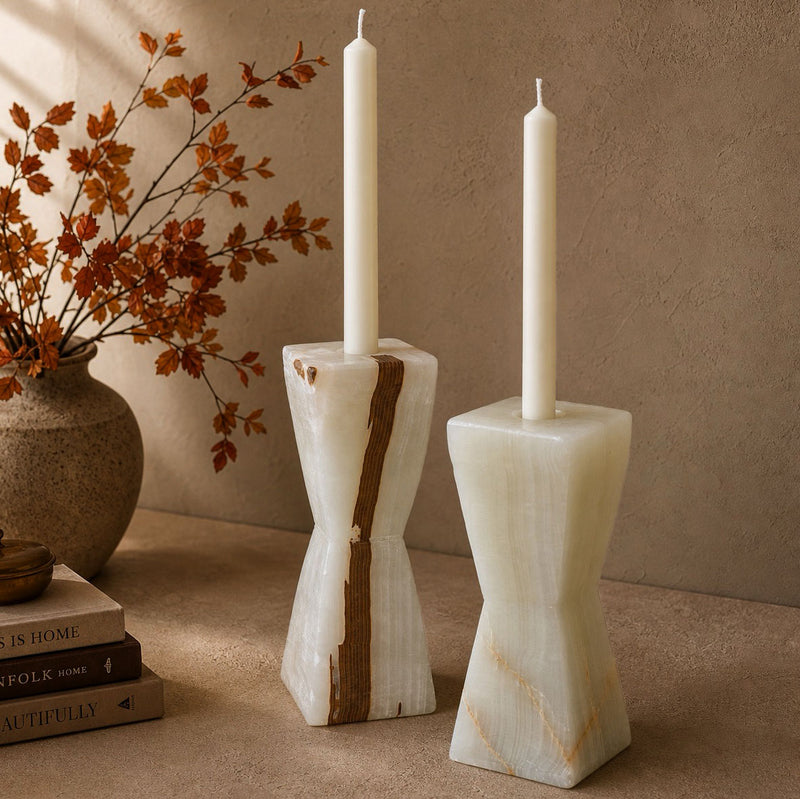 Pair of ivory onyx hourglass candle holders with white taper candles, styled beside a ceramic vase of dried autumn branches and stacked books.