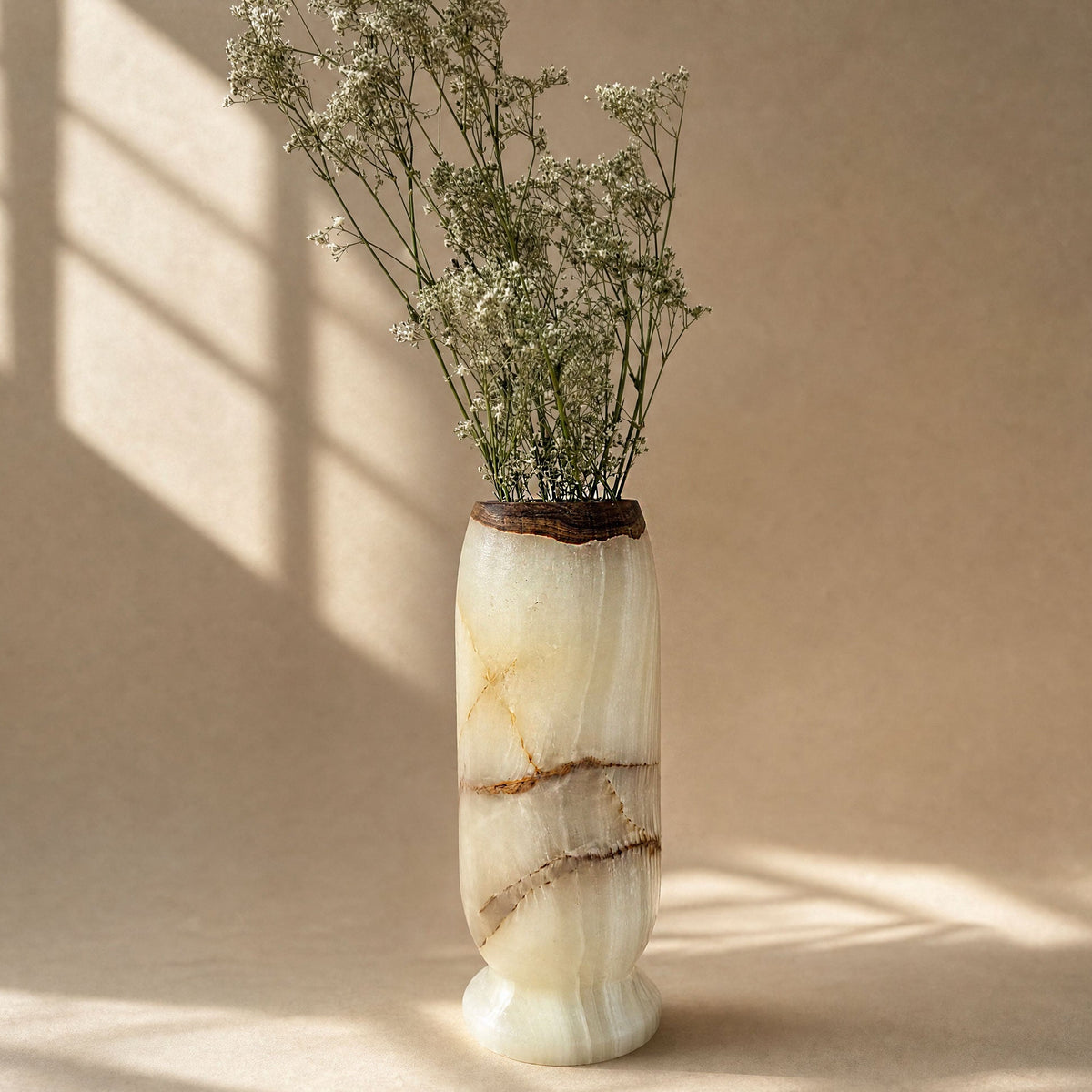 A tall cylindrical vase made of cream and beige marbled onyx stone with brown veining and a dark natural wood top, holding dried white flowers, set against a warm beige background with soft window-shadow light.