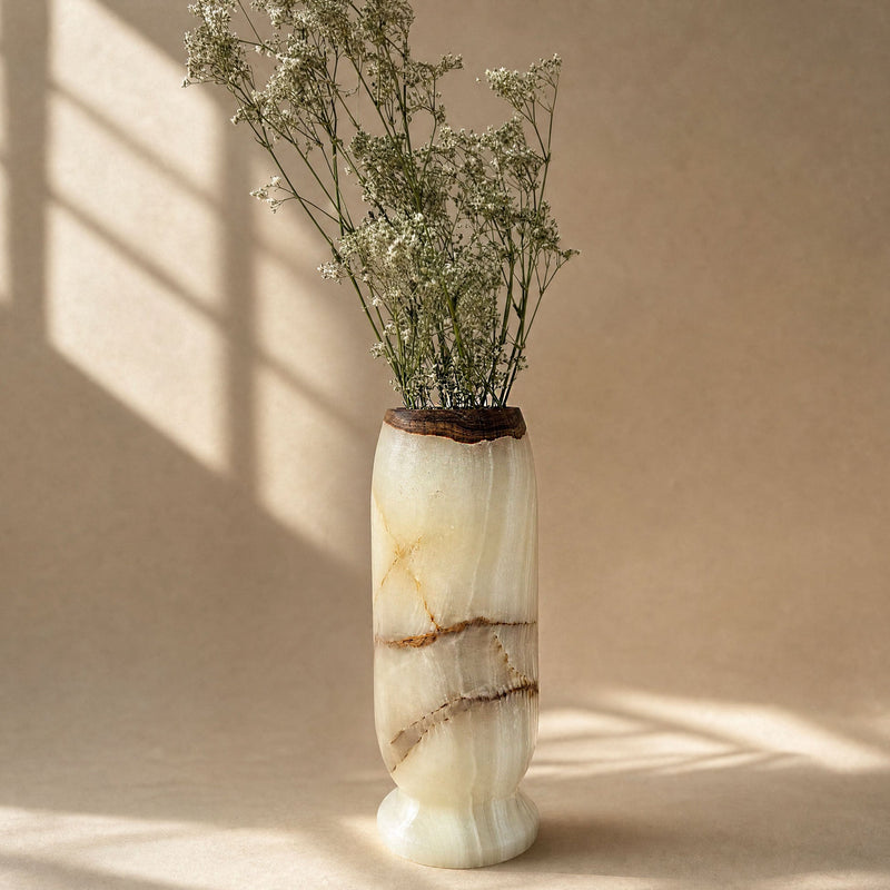 A tall cylindrical vase made of cream and beige marbled onyx stone with brown veining and a dark natural wood top, holding dried white flowers, set against a warm beige background with soft window-shadow light.