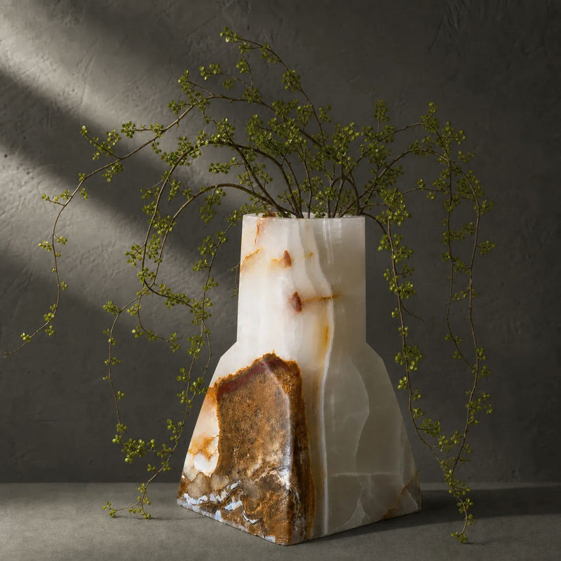 Ivory and cognac onyx bud vase holding trailing green stems, a crystalline cognac drift banding across its tapered base, against a dark plaster wall in raking light.