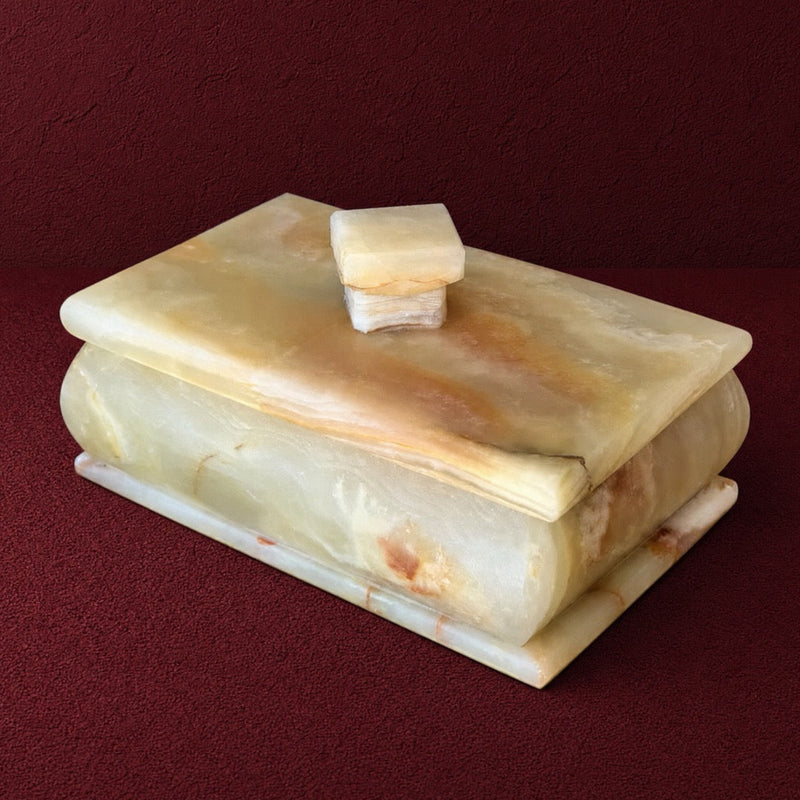 A rectangular lidded stone box made of pale cream and tan onyx with natural veining, photographed on a dark red textured background.