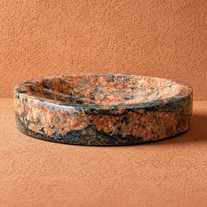 A round stone bowl dish with a mottled granite pattern in orange, brown, and dark gray, set against a textured terracotta background.