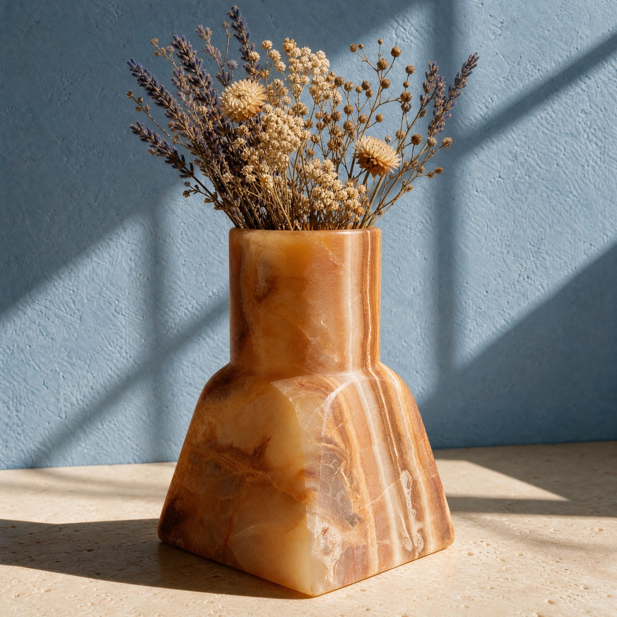 A sculptural terracotta-colored stone vase filled with dried flowers, set on a light stone surface against a textured blue wall with soft window shadows.