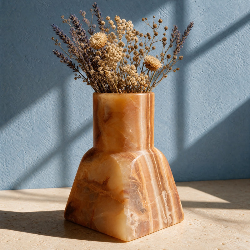 A sculptural terracotta-colored stone vase filled with dried flowers, set on a light stone surface against a textured blue wall with soft window shadows.