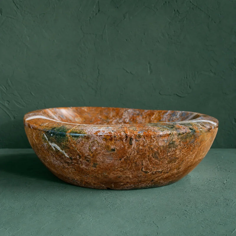 A round stone vessel sink bowl in a warm brown and orange marbled pattern with natural white and dark green veining, set against a dark green textured background.