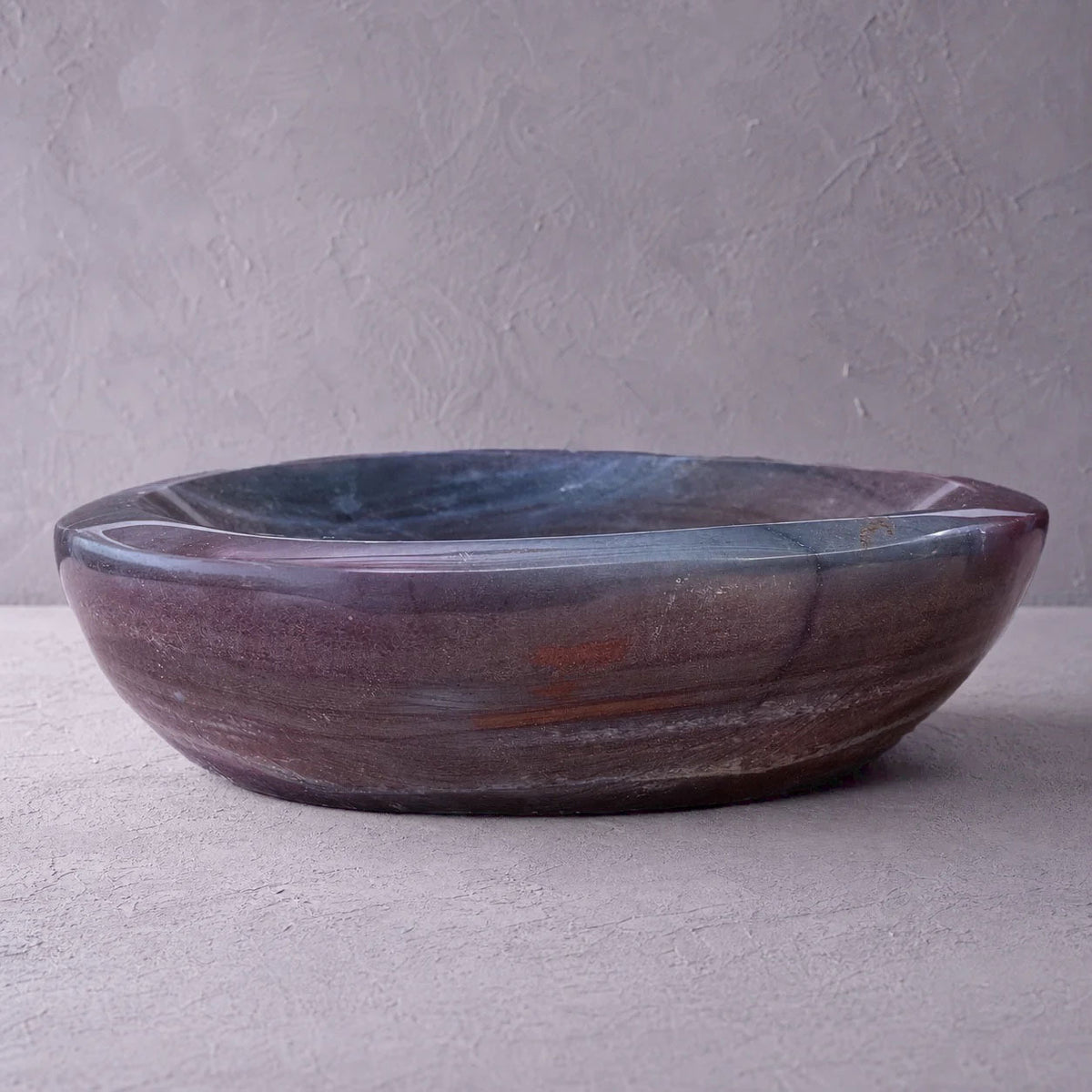 A round stone vessel bowl with a marbled purple, brown, and blue-gray finish, set against a textured gray concrete background.
