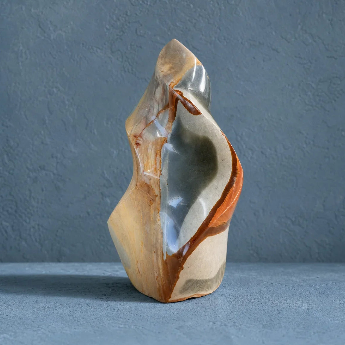 A polished abstract stone sculpture with flowing organic contours, featuring a mix of cream, tan, gray, and rust-orange tones, set against a textured blue-gray background.
