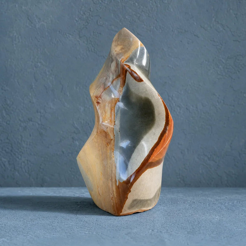 A polished abstract stone sculpture with flowing organic contours, featuring a mix of cream, tan, gray, and rust-orange tones, set against a textured blue-gray background.