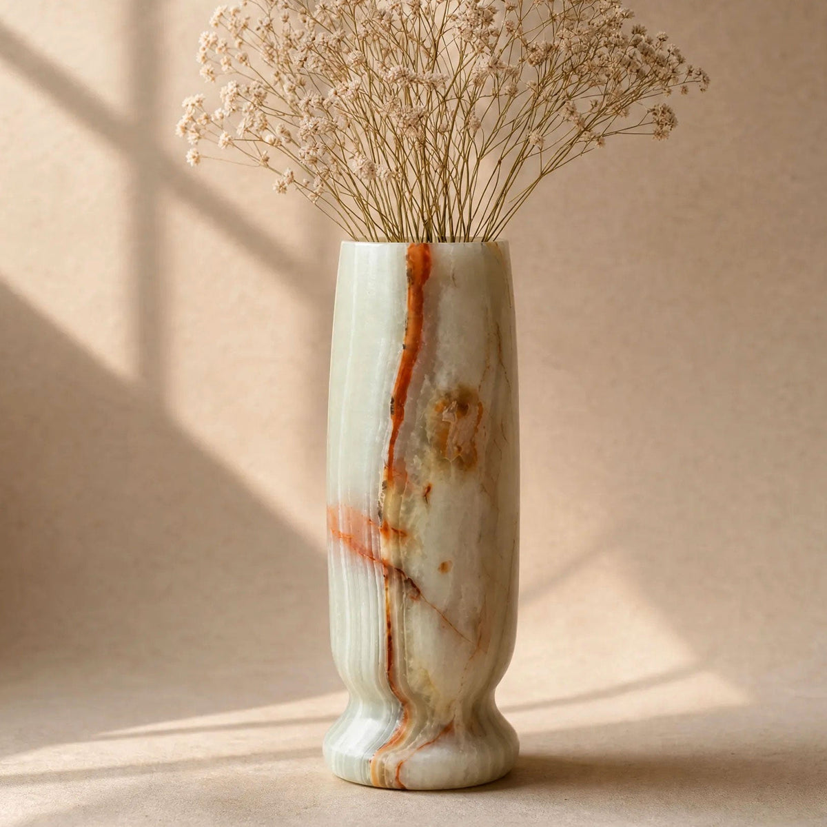 A tall cylindrical marble vase with a flared base, featuring natural white, beige, and rust-orange veining, filled with dried baby's breath flowers against a warm beige background with soft window shadow patterns.