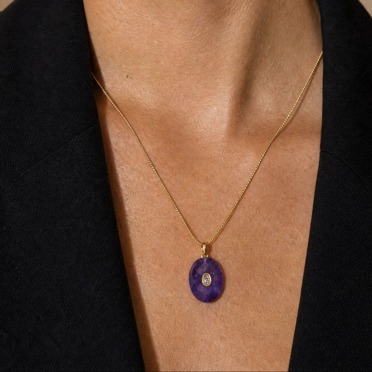 Close-up of a person wearing a gold necklace with a purple pendant against a neutral background